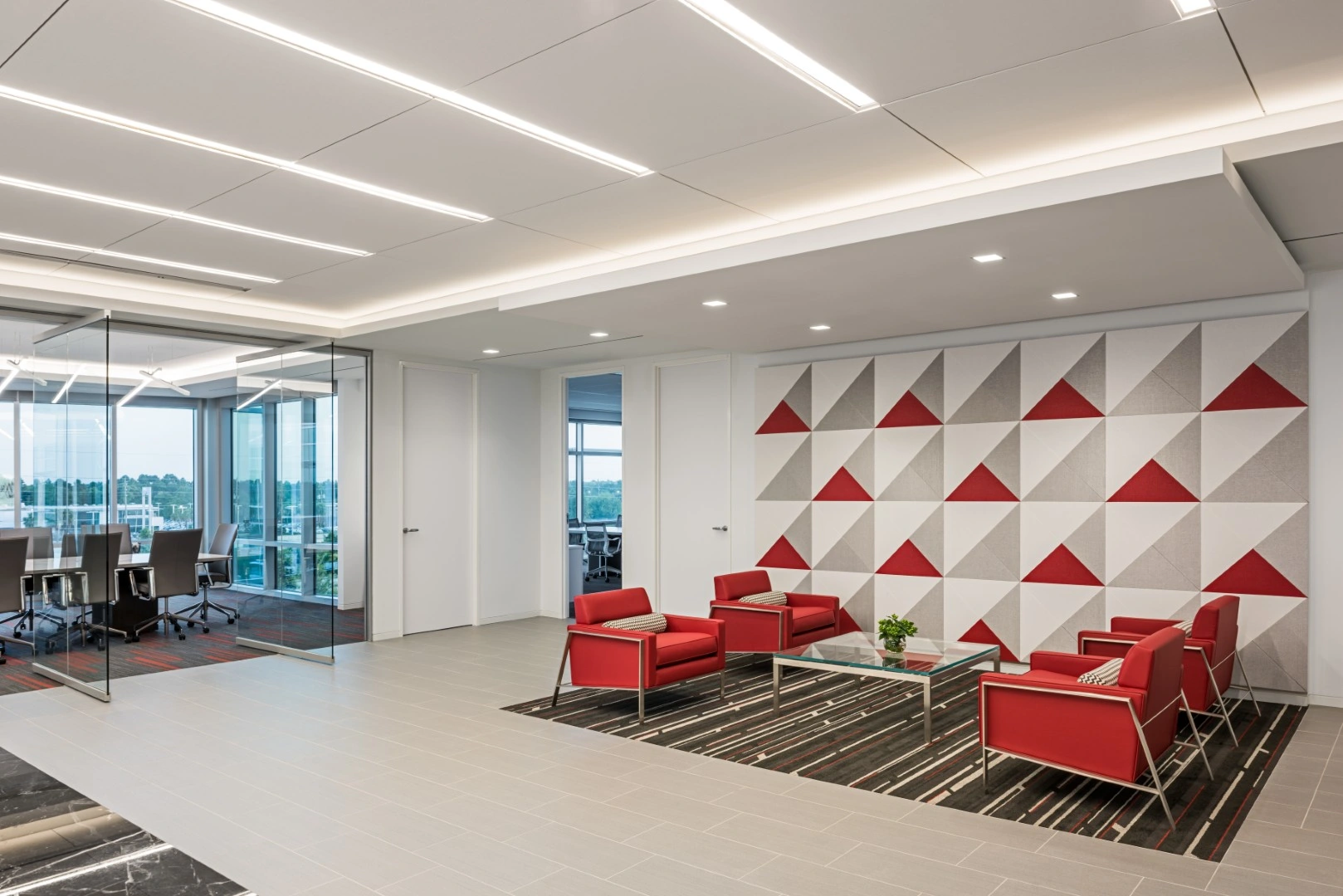 A modern office lounge with red chairs, glass walls, and a geometric wall design of red, gray, and white fabric.