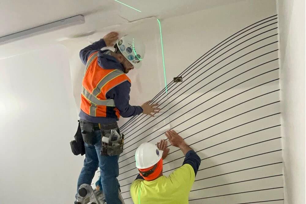 Two workers, one on a ladder and one below, use a green laser level to align lines on a wall for construction or painting.