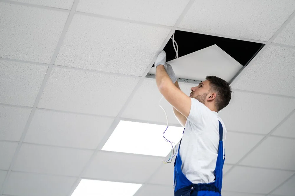 A worker in gloves installs a ceiling panel, revealing a dark space above a white ceiling with grid tiles and overhead lighting.