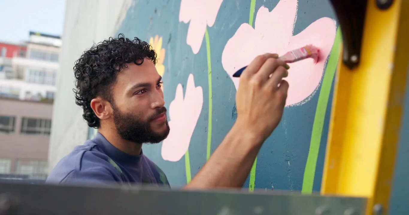 A person with curly hair paints pink flowers on a vibrant wall, showing creativity and artistic expression.